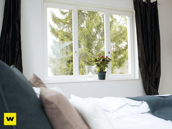 Helles Schlafzimmer mit großen Fenstern, Bett, Kissen und Topfpflanze am Fenster.